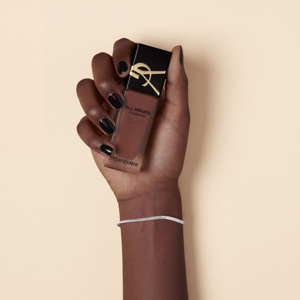A hand with dark nail polish holding a bottle of YSL All Hours Foundation against a neutral background.
