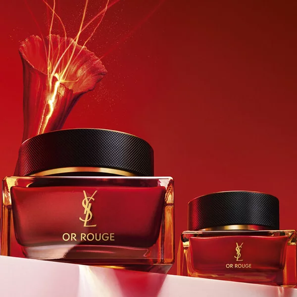A striking display of two luxurious red skincare jars, featuring the YSL Or Rouge branding, set against a vibrant red background, with golden sparks emerging from the larger jar.