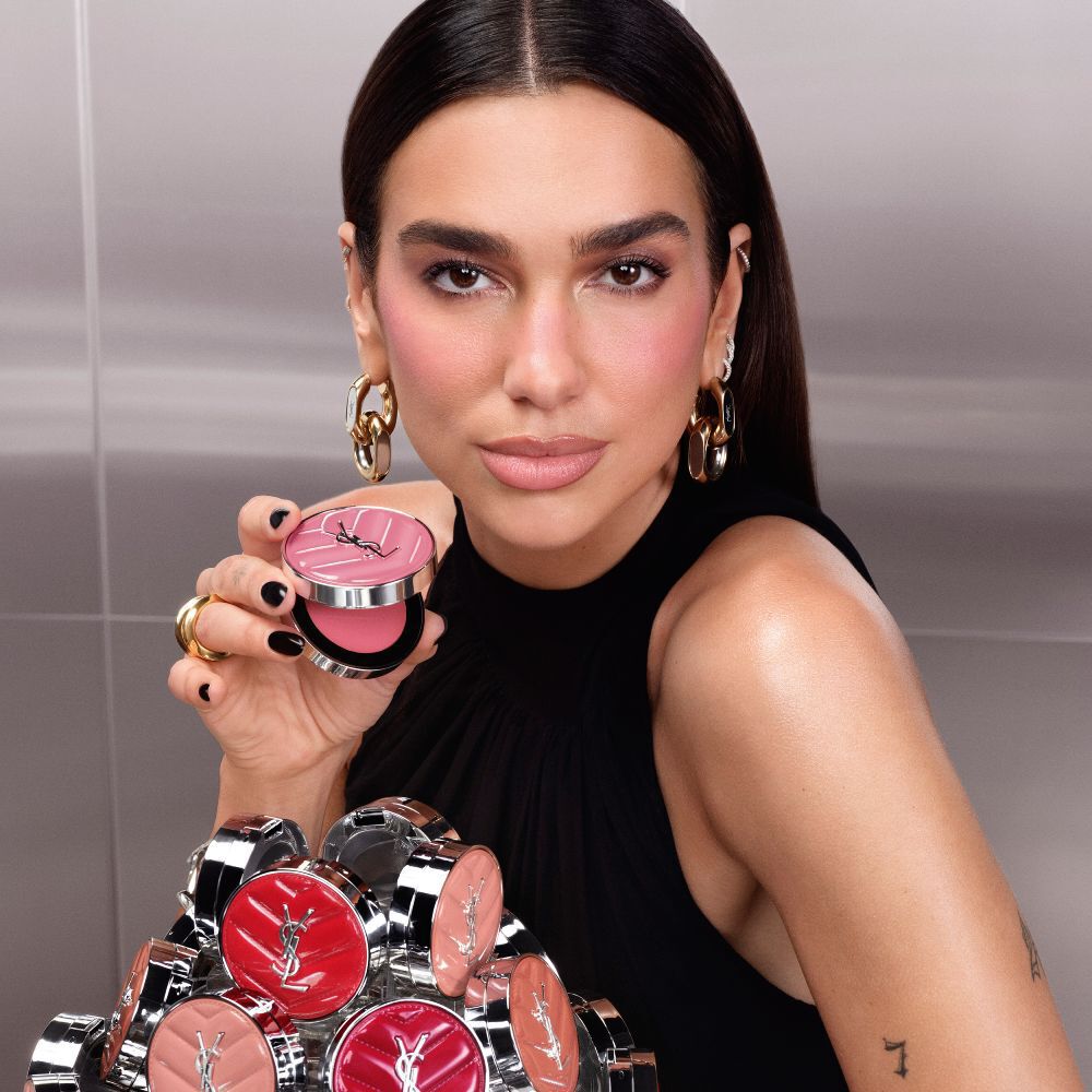 Dua Lipa tient un boitier ouvert de YSL Make Me Blush Lifted Glow Cream Blush, une collection des nouvelles teintes etalee devant elle.