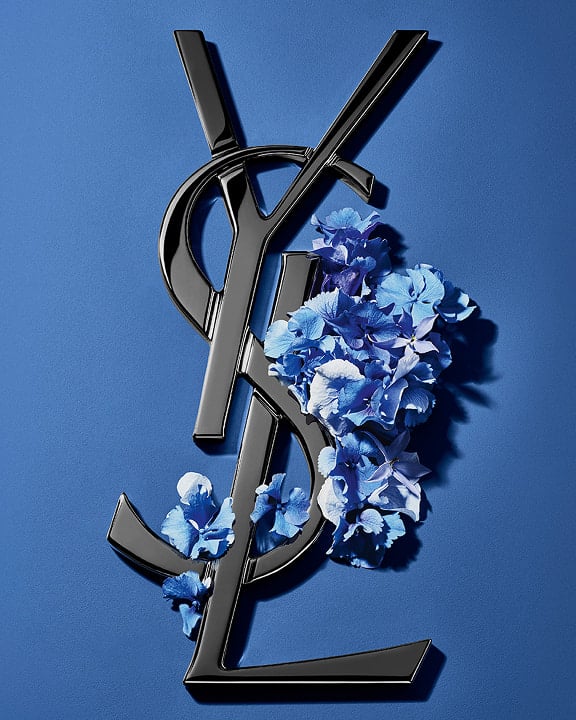 Schwarzes YSL Cassandre-Logo, verziert mit leuchtend blauen Frühlingsblumen, vor einem ruhigen blauen Hintergrund.