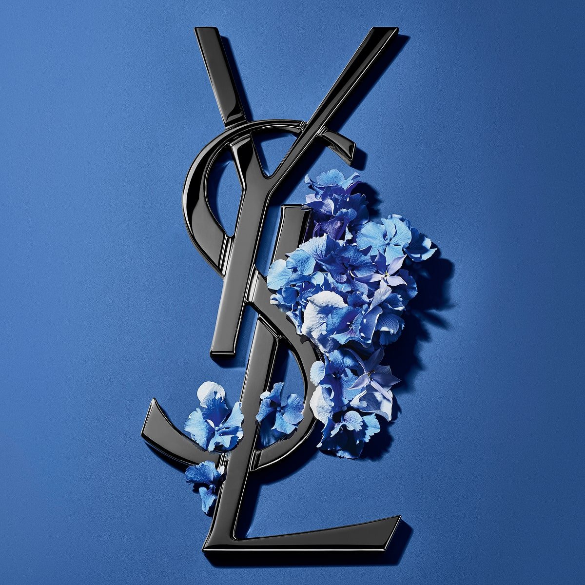 Schwarzes YSL Cassandre-Logo, verziert mit leuchtend blauen Frühlingsblumen, vor einem ruhigen blauen Hintergrund.