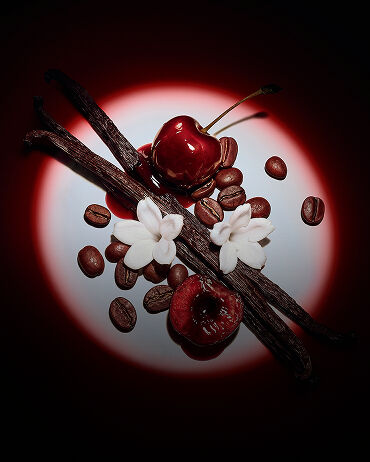 Nahaufnahme saftiger roter Kirschen, Jasminblüten, Vanilleschoten und Kaffeebohnen, dramatisch von einem Spotlicht für Black Opium Eau de Parfum Over Red in Szene gesetzt.