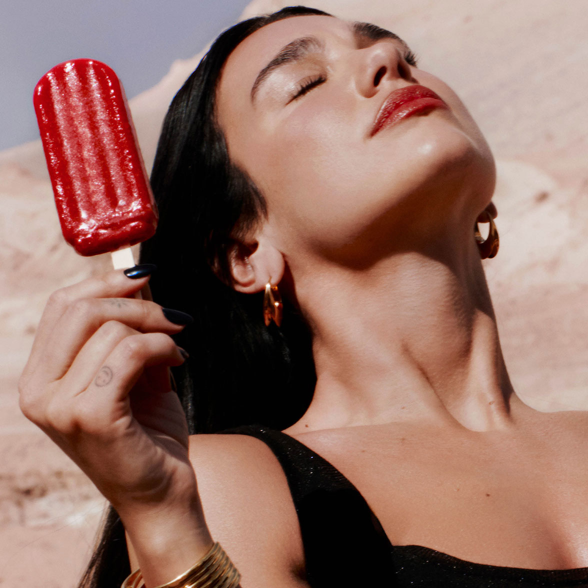 Eine Nahaufnahme von Dua Lipa mit geschlossenen Augen in der Sonne, die ein rotes Wassereis hält.